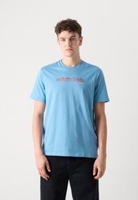 Šviesiai mėlynas medvilninis marškinėlis su raudonu "Calvin Klein" logotipu ant krūtinės, apvaliu kaklu ir trumpomis rankovėmis.