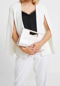 Metallinen hopeanvärinen clutch-laukku, jossa on jämäkkä muoto, suorakulmainen aukko kahvassa ja hillitty hohde. Yhdistetty valkoiseen viittaan.