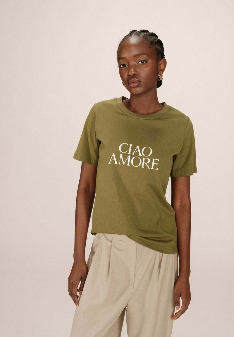 T-shirt en coton vert olive avec "CIAO AMORE" en texte blanc, col rond, manches courtes, associé à un pantalon beige clair. Texture lisse.