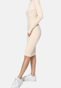 Robe en tricot beige à manches longues avec texture côtelée, silhouette ajustée, longueur genoux, associée à des baskets blanches.