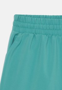 Shorts de sport turquoise avec une taille élastique froncée et une poche latérale, fabriqués en tissu lisse et léger.