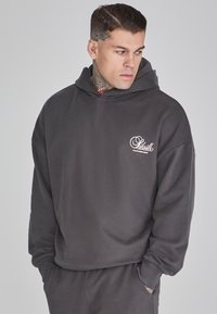 Felpa oversize grigia scura con tasca frontale, cappuccio classico e logo tono su tono sul petto. Tessuto morbido con una texture liscia.