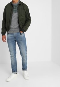 Man med grön bomberjacka, gråmönstrad tröja, ljusblåjeans med uppvikta byxben och vita sneakers, stående med en hand i fickan.