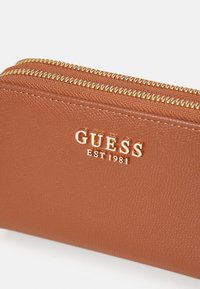 Rudas tekstūruotas odinis piniginė su auksine užtrauktuko sistema ir išspaustu "GUESS EST 1981" logotipu priekyje. Kompaktiškas dizainas su praktišku uždarymu.