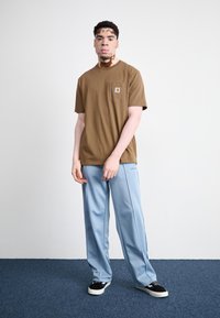 T-shirt de algodão castanho com detalhe de bolso, combinado com calças azuis claras. O modelo usa ténis pretos e brancos sobre um tapete azul.