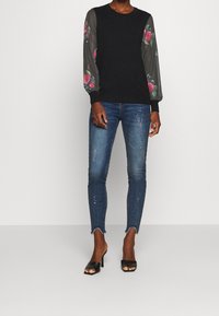 Kvinna i svart topp med genomskinliga blommönstrade ärmar, slitna blå skinny jeans med råa fållar, och svarta öppna högklackade mule-skor.
