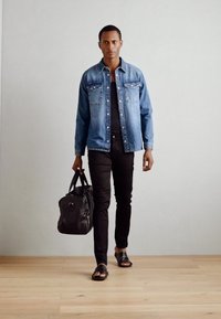 Kék farmerdzseki fekete pólón, fekete slim-fit nadrágban és szandálban. Fekete sporttáska vállpánttal.