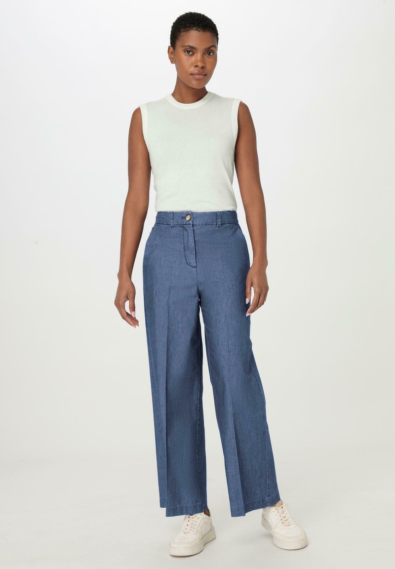 Jasnoszara bezrękawnik sparowany z szerokimi, ciemnoniebieskimi dżinsami z guzikiem i fałdami z przodu, uzupełniony białymi sneakersami.