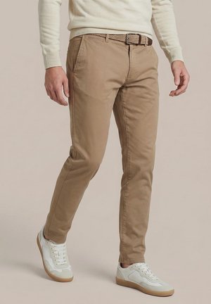 Man die beige chino's draagt, een geweven riem, witte sneakers en een crème lange mouwen trui, staand met één been iets naar voren.