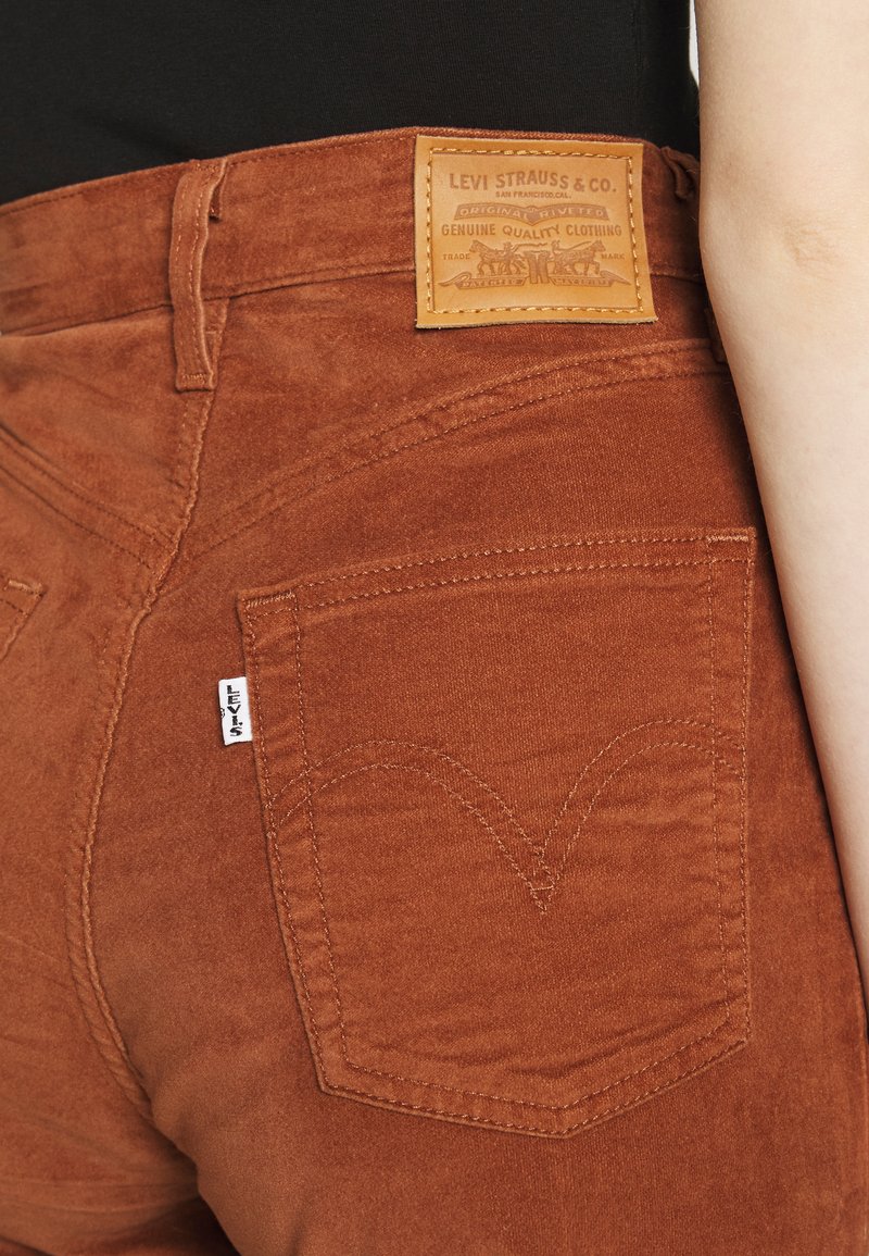 Rostfarbene Wildlederjeans mit einem Leder-Label-Patch an der Taille, klassischen Gesäßtaschen und sichtbaren Nähdetails.