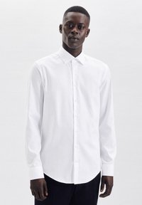 Camicia bianca a maniche lunghe realizzata in tessuto morbido, con colletto classico e chiusura con bottoni, orlo arrotondato e maniche aderenti.