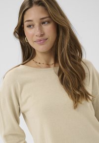 Pull en tricot beige avec une coupe large et un col large. Il présente des manches longues et une texture douce, associé à un simple collier de perles.