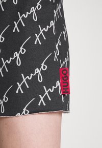 Shorts noirs avec un motif manuscrit blanc "Hugo" et une étiquette en tissu rouge affichant "HUGO". Fabriqué en coton texturé.