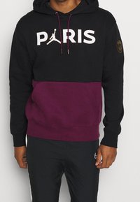 Sweat à capuche noir avec une section inférieure violette. Présente "PARIS" et un logo Jumpman en blanc. Comprend une poche avant et des poignets élastiques.
