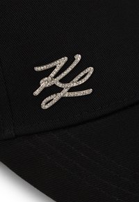 Căciulă neagră dintr-un material texturat, cu un emblema strălucitoare de argint stilizată sub forma literei "K" pe față, sporind contrastul vizual.