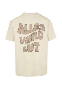 Cremefarvet bomulds-t-shirt med fed, brun tekst på ryggen, der står "ALLES WIRD GUT" i en legesyg, rund skrift.