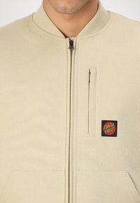 Beige bomber dzseki bordázott gallérral, cipzáras záródással és oldalsó zsebekkel. A bal felső oldalon fekete és piros "Santa Cruz" logójal ellátott folt található.