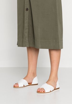 Białe płaskie sandały z krzyżującymi się paskami, wykonane ze skóry, z odkrytymi palcami i brązową podeszwą, prezentowane na jasnej podłodze.