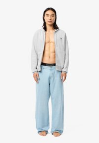 Szürke cipzáras kapucnis pulóver oldalsó zsebekkel, világoskék, bő szárú farmernadrággal párosítva. Fekete derékrész "LACOSTE" logóval. A modell mezítláb áll.