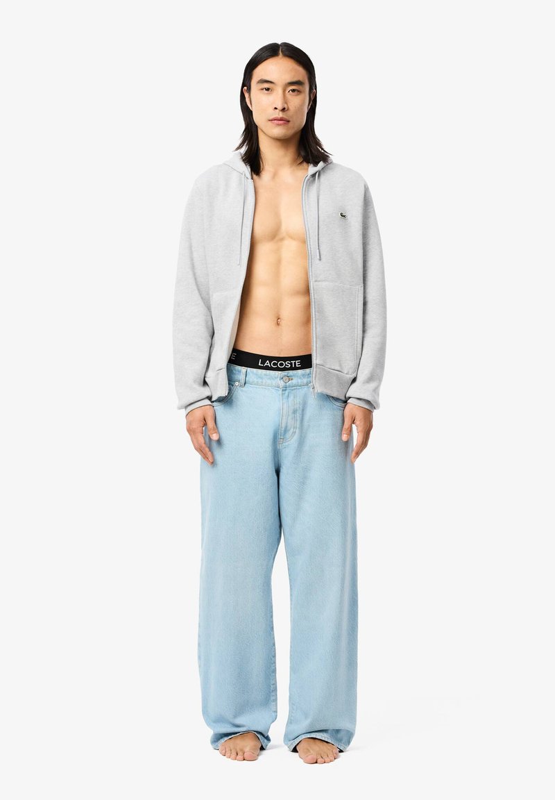 Haut de sweat à capuche gris zippé avec poches latérales, associé à un jean large bleu clair. Ceinture noire avec le logo "LACOSTE". Le modèle est pieds nus.