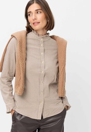 Frau trägt ein kariertes Hemd mit Knopfleiste, Rüschenkragen und Manschetten, einen braunen Strickpullover über den Schultern und dunkle Hosen, schaut seitlich.