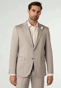 Man wearing a beige suit with white shirt and a small white flower pin on the left lapel, standing against a plain light background.