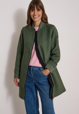 Femme souriante portant un long manteau vert zippé, un top rose et un jean bleu, une main dans la poche sur un fond clair uni.