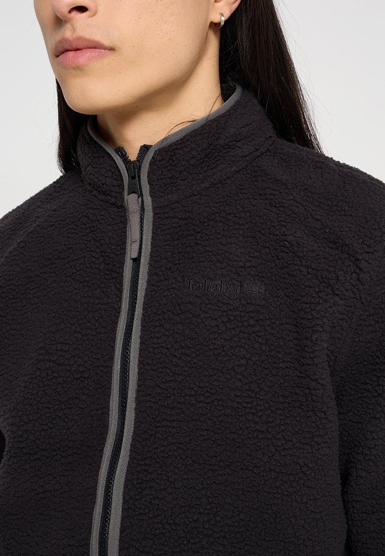 Giacca in pile nera con colletto alto, dotata di chiusura a zip e accenti grigi. Il tessuto ha una finitura testurizzata e un dettaglio con logo.