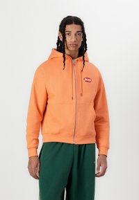 Orange zip-up hoodie made of cotton blend, featuring a front pocket and logo patch. Model wears it with green sweatpants.