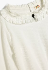 Blusa bianca a manica lunga con un motivo texture, caratterizzata da un colletto arricciato e un nastro nero di accento al colletto.