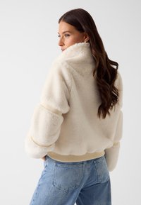 Puszysta kremowa kurtka z wysokim kołnierzem i elastycznymi mankietami. Charakteryzuje się miękką fakturą i prążkowanym obszyciem u dołu, zestawiona z niebieskimi dżinsami.