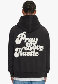 Schwarzer Hoodie mit großen weißen Schriftzügen auf dem Rücken, die "Pray Love Hustle" lesen. Das Material sieht weich aus und hat eine normale Passform mit Kapuze.