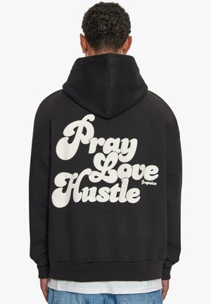 Black hoodie featuring large white text on the back that reads "Pray Love Hustle." Material appears soft with a standard fit and hood.