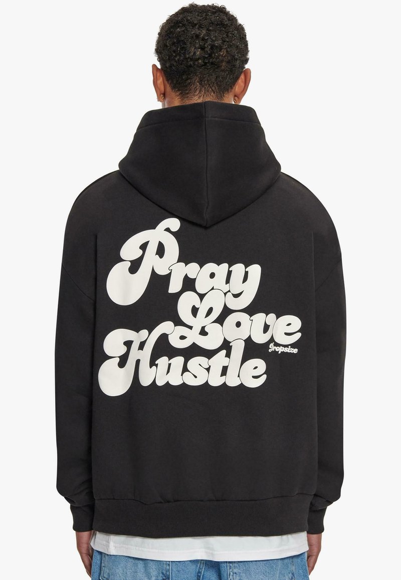 Schwarzer Hoodie mit großen weißen Schriftzügen auf dem Rücken, die "Pray Love Hustle" lesen. Das Material sieht weich aus und hat eine normale Passform mit Kapuze.