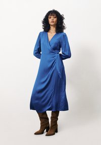 Blaue Satin-Wickelkleid mit langen Puffärmeln und einem Taillengürtel. Knielang, mit glatter Textur, ergänzt durch knielange braune Wildlederstiefel.