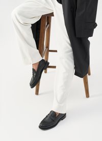 Mustad nahast loafers, millel on sile viimistlus ja õmblustega detailid, koos valgete pükstega, puidust toolil.