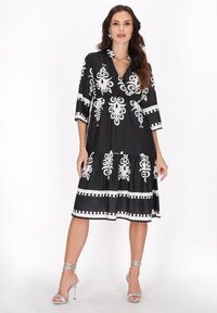 Rochie neagră cu modele florale albe, mâneci trei sferturi și fustă în straturi. Prezintă un decolteu în V și detalii decorative la tiv.
