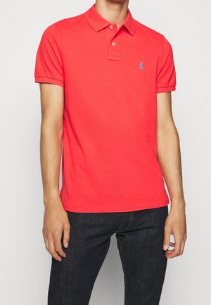 Homme portant un polo rouge vif à manches courtes avec un petit logo bleu sur la poitrine et un jean bleu foncé, debout devant un fond uni.