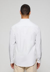Camicia bianca a manica lunga con sottili righe, colletto classico, polsini con bottoni e dettagli sulla spalla posteriore, realizzata in tessuto leggero.
