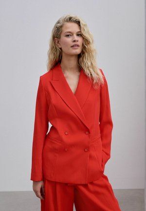Vrouw met lang krullend blond haar, gekleed in een rood dubbelgeknoopt jasje en bijpassende broek, staand tegen een eenvoudige lichte achtergrond.