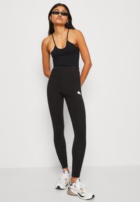 Leggings atléticos pretos com cintura alta, tecido elástico, risca lateral branca e pequeno detalhe de logótipo. Combinados com uma t-shirt justa preta e ténis.