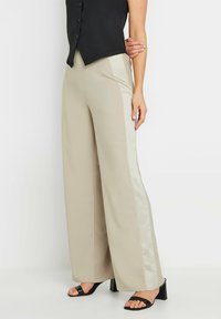 Pantalons larges beige avec une bande satinée sur le côté ; texture lisse ; portés avec un top sans manches noir et des sandales à talons noires.