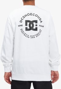 Person wearing a white sweatshirt with black DC Shoes logo and circular text on the back reading "DCSHOECOUSA Nine Four Athletic Skateboard Footwear."