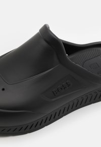 Shoe preto de fácil calçar, feito de borracha lisa com uma sola texturizada. Apresenta um logótipo da marca visível e um design elegante sem decorações adicionais.