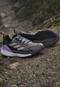 Buty do biegania terenowego z wytrzymałymi czarnymi podeszwami, szarymi cholewkami, fioletowymi piętami i jasnoniebieskimi sznurówkami umieszczone na skalistym terenie na zewnątrz.