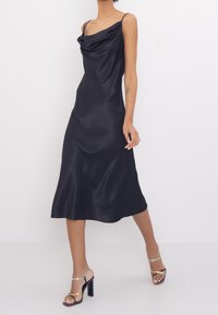 Black satin dress with a cowl neckline, thin straps, and a flowing silhouette, complemented by strappy high-heel sandals.