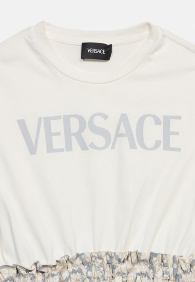 Εκρού τοπ Versace με μεγάλο γκρι λογότυπο, στρογγυλή λαιμόκοψη και τελείωμα με βολάν σε ανθικό μοτίβο.