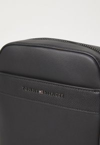 Svart syntetmaterialväska med ett texturerat band. Har ett dragkedjestängning och en metallogo "TOMMY HILFIGER" på framsidan.