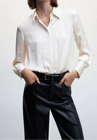 Chemise blanche à manches longues avec col, associée à un pantalon en cuir noir taille haute et une ceinture noire. Texture lisse, coupe ajustée.