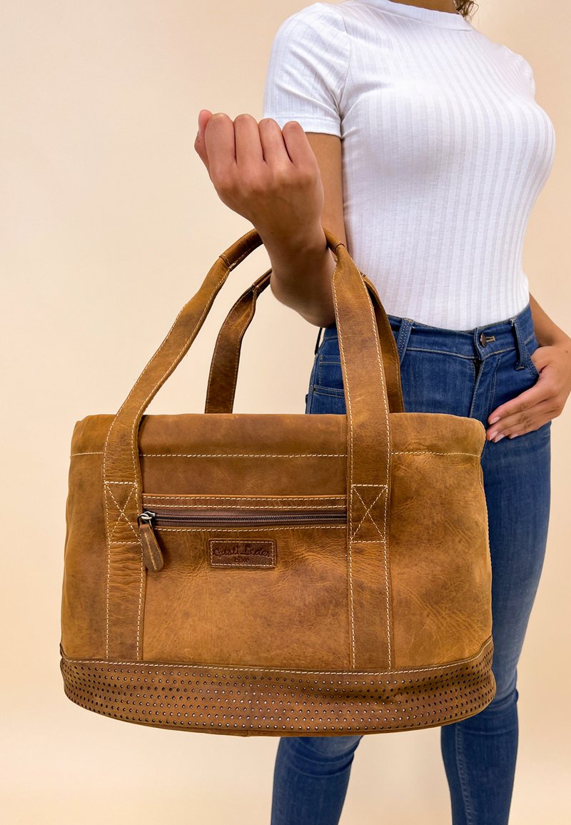 Bruine leren tote bag met twee handvatten, ritsvak, decoratieve geperforeerde bodem en contrasterende stiksels. Afmetingen niet gespecificeerd.